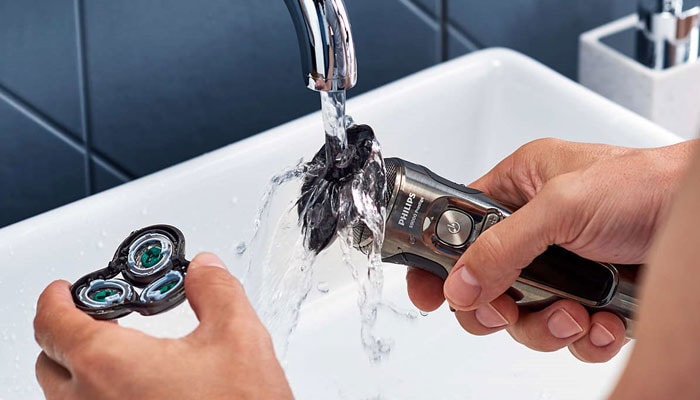 Close-up of person holding two parts of an electric shaver in both hands under a water running tap.