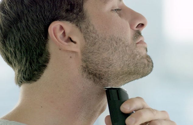 Side profile of a man with short beard shaving his neck with an electric shaver.