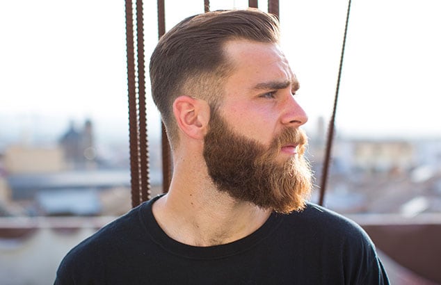 Side profile of man with a long, brown beard and short, brown hair with a city out-of-focus in the background.