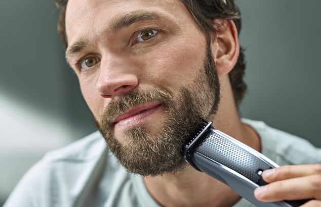A bearded man with brown hair using an electric shaver.