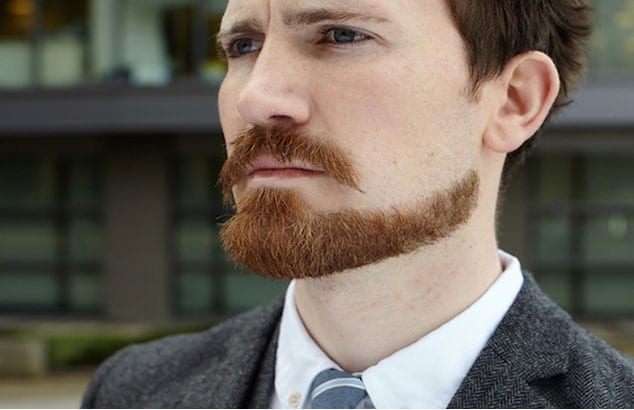 Man with an accurately cut ginger chin strap beard in a dark grey suit is looking into the distance.