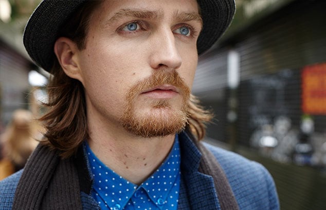 : Man with shoulder long hair and Henri quatre chin beard wearing a blue shirt, a suit jacket, and a hat.