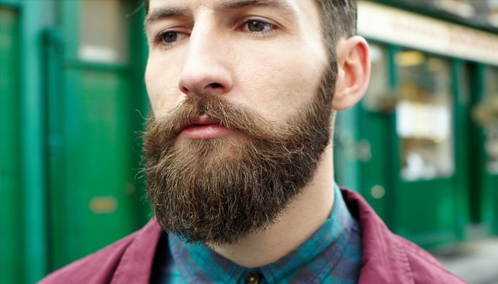 Man with full brown beard and moustache standing in a street wearing checked shirt and maroon jacket.