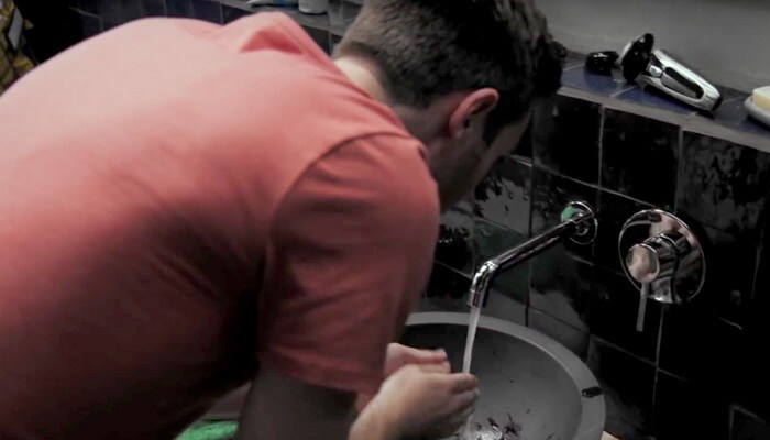 The back of a man in red t-shirt as he puts his hands under running water from a tap in a dark bathroom.